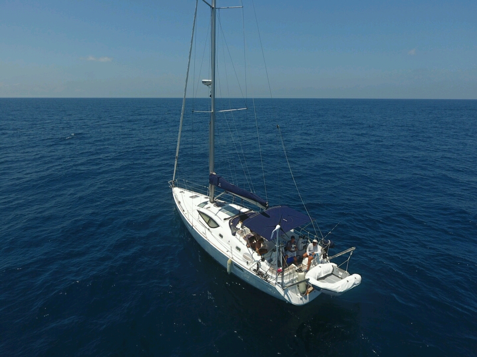 Elevated drone view of a white monohull sailboat anchored in deep blue open sea, sails furled, blue bimini over the cockpit, a few passengers aft and an inflatable dinghy hanging off the stern.
