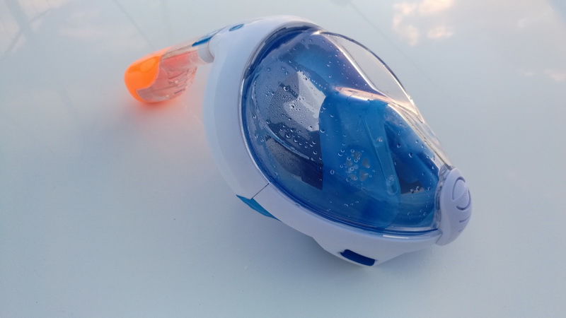Close-up product-style photo of a full-face snorkel mask, white and blue with orange snorkel tip, covered in water droplets, resting on a white deck surface.
