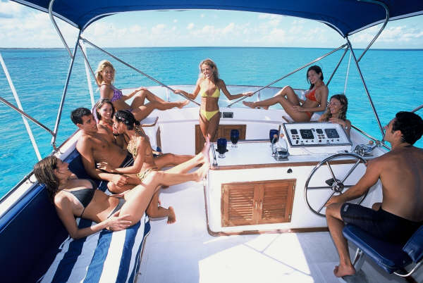 Group of young people in swimwear lounging on the flybridge of a motor yacht under a blue bimini, one at the helm wheel, striped cushions, turquoise sea stretching behind.