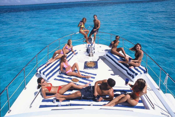 Ten young guests in swimwear sunbathing and chatting on the bow of a white motor yacht with blue-striped sunpads, snack trays out, anchored on brilliant turquoise shallow water.