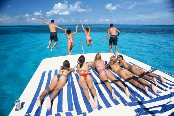 Four people jumping off a yacht's bow into turquoise water while six others sunbathe face-down on blue-and-white striped towels on the foredeck, sunny Caribbean day.