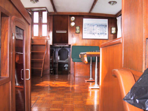 Classic motor yacht interior with varnished wood paneling and parquet floor, green-cushioned bench seating, small table, ladder to deck and framed print on the wall.