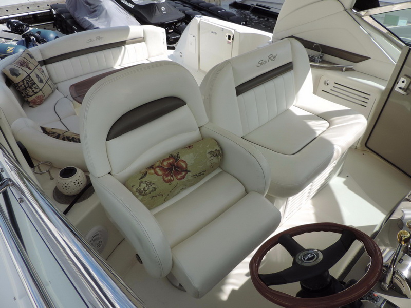 Close view of Sea Ray helm and companion bucket seats in white leather with flower-print lumbar pillow, wood-rimmed steering wheel, curved rear lounge with sea-themed pillows behind.
