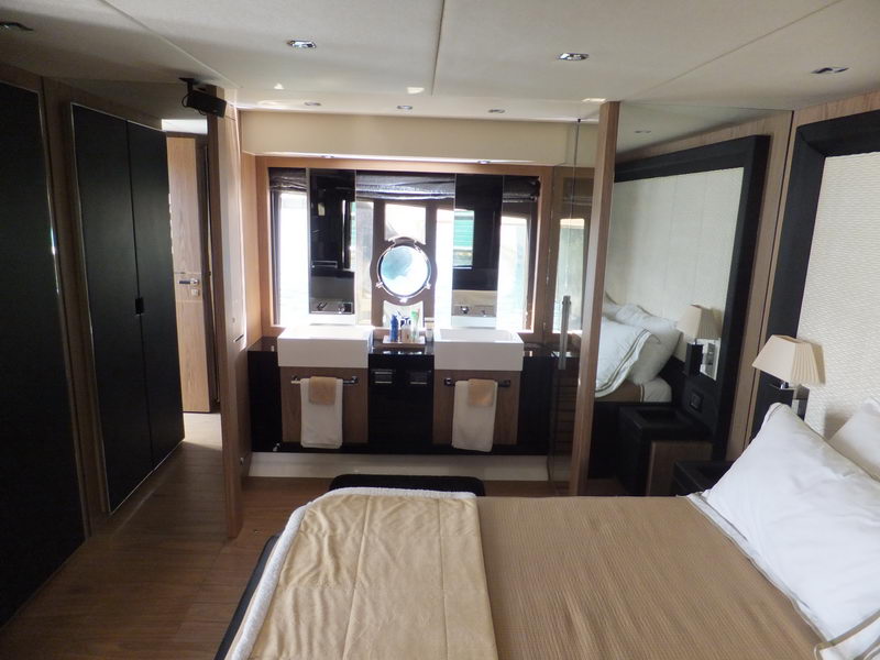 Master cabin interior with double bed in gold bedspread, dark wood joinery, twin white vessel sinks with towels, round porthole mirror, mirrored wardrobe panels and warm ceiling lighting.