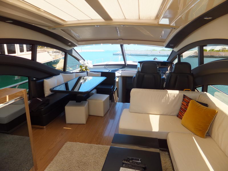 Sport yacht upper salon with two black helm seats, glass-top dining table with black bench, white sectional sofa with yellow and striped cushions, wood floor, marina water visible through open sides.