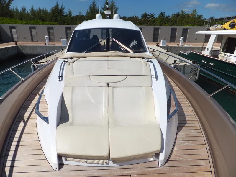 Bow of a white sport yacht with large cream sunpad cushions and stainless rails, seen head-on from the dock, tan canvas windshield cover, green trees and marina wall behind, neighboring boats to the right.