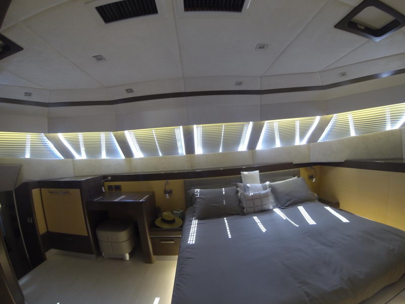 Master cabin of a catamaran with king bed in gray linens, sunlight streaming through slatted blinds along a panoramic window band, vanity desk with stool, straw hat on nightstand, cream and yellow paneling.