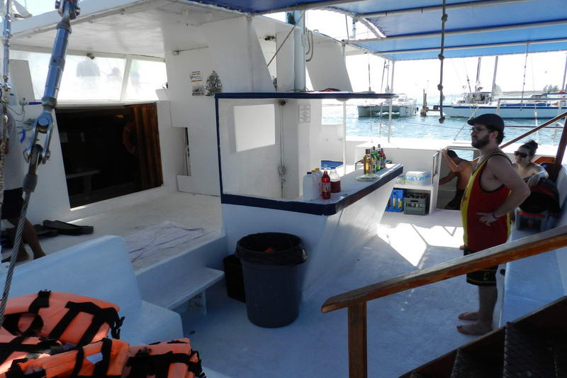 Catamaran deck with an onboard bar counter stocked with bottles, a pile of orange life jackets in the foreground, a trash bin, and a man in a tank top photographing the marina with his phone.