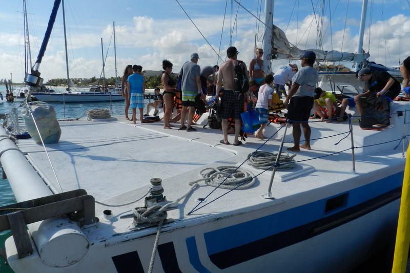 Close-up of a catamaran's foredeck at the marina with several passengers standing (some in swimwear, one with a backpack), coiled ropes on deck, and other sailboats moored nearby.
