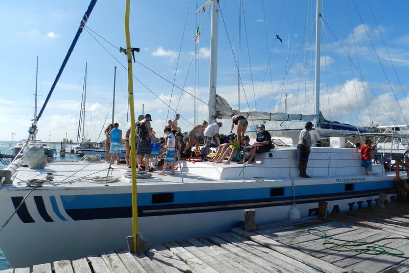 Catamaran docked at a wooden pier with numerous passengers, including children, gathered on deck getting ready for departure, other masts visible in the marina.