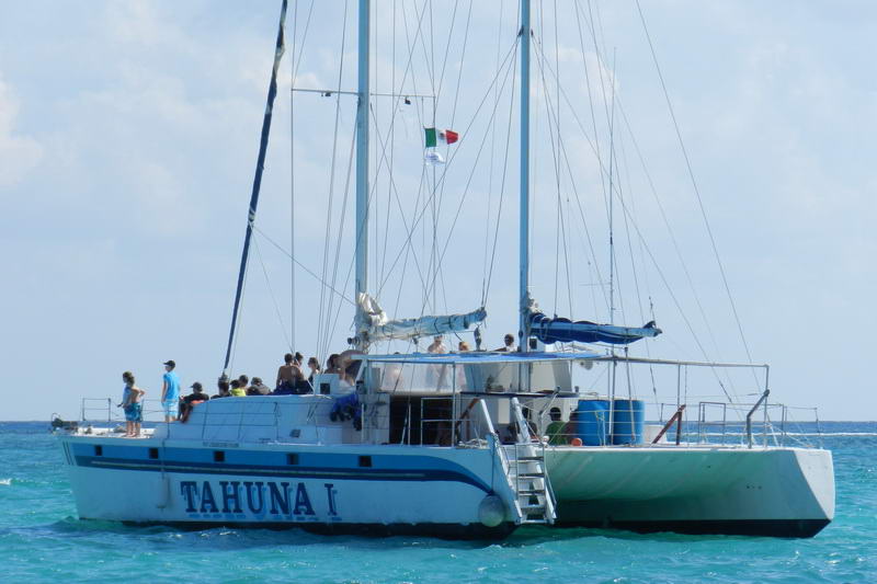Sailing catamaran named 'Tahuna I' anchored on calm turquoise water with passengers standing on deck under a clear sky, a Mexican flag flying from the mast.